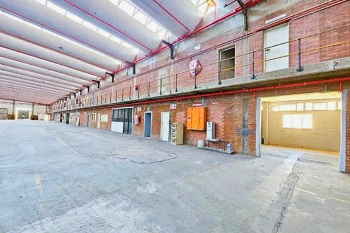 Expansive industrial warehouse space featuring high ceilings with skylights, exposed brick walls, concrete flooring, and a mezzanine level with metal railings.