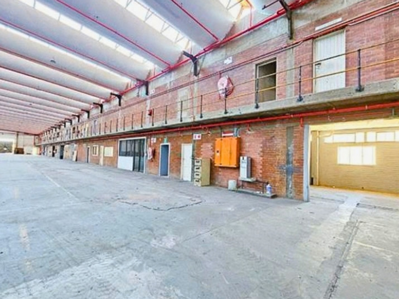 Expansive industrial warehouse space featuring high ceilings with skylights, exposed brick walls, concrete flooring, and a mezzanine level with metal railings.