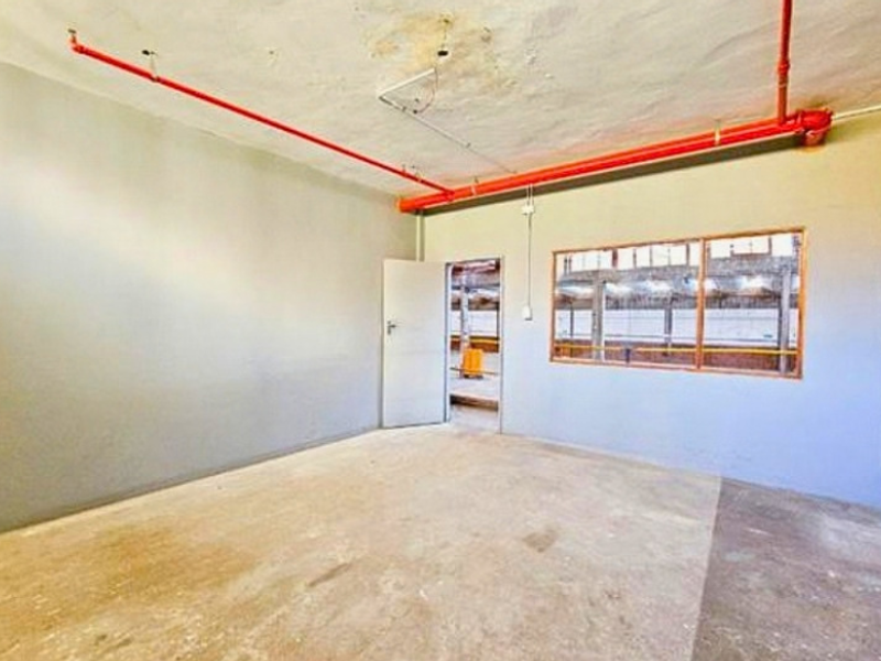 An empty industrial utility space featuring exposed fire sprinkler piping, concrete flooring, and a large window overlooking an adjacent warehouse area.
