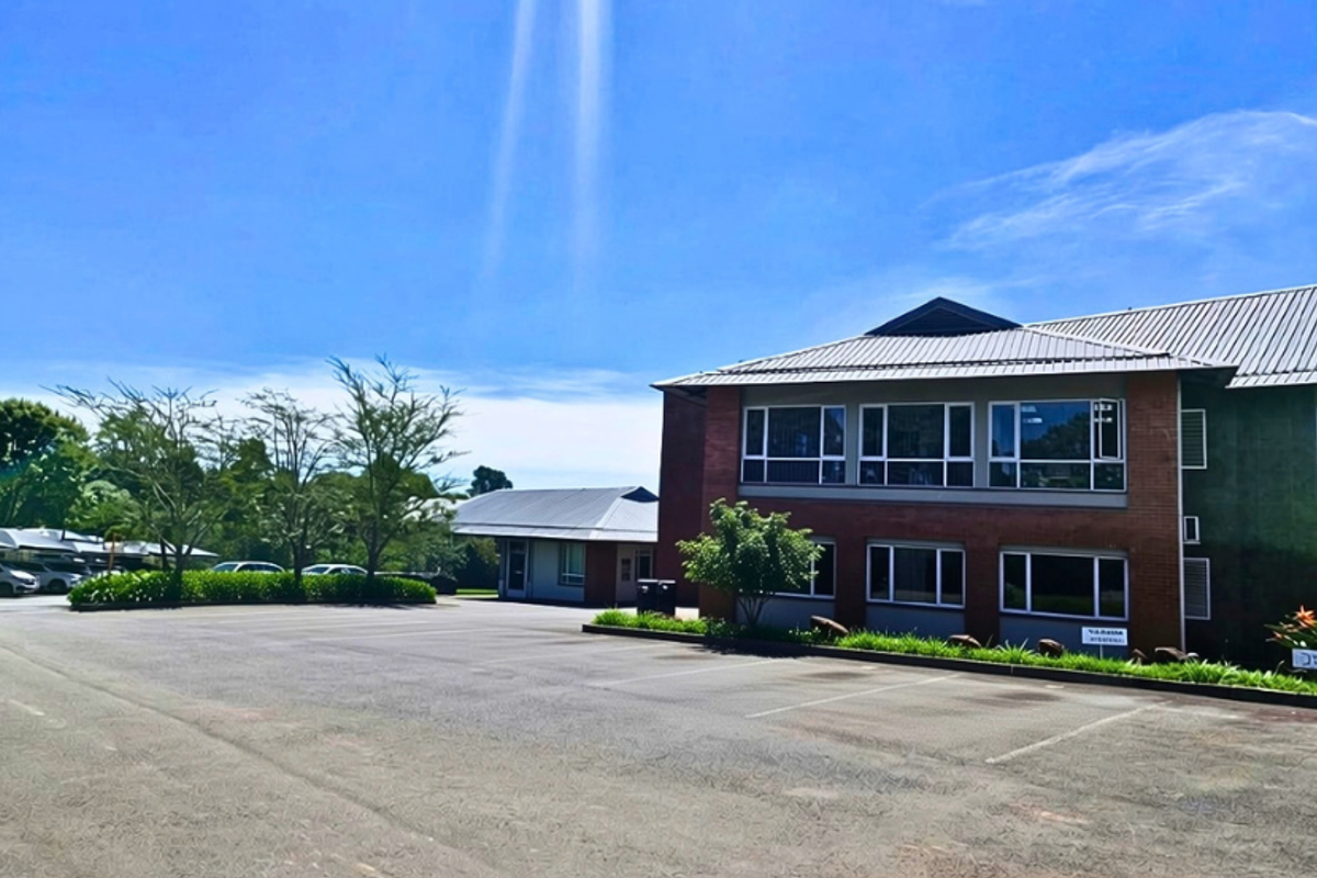 A professional office building exterior featuring a brick facade, metal roofing, large windows, and an expansive asphalt parking area.