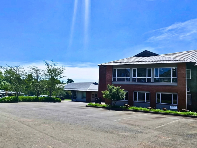 A professional office building exterior featuring a brick facade, metal roofing, large windows, and an expansive asphalt parking area.