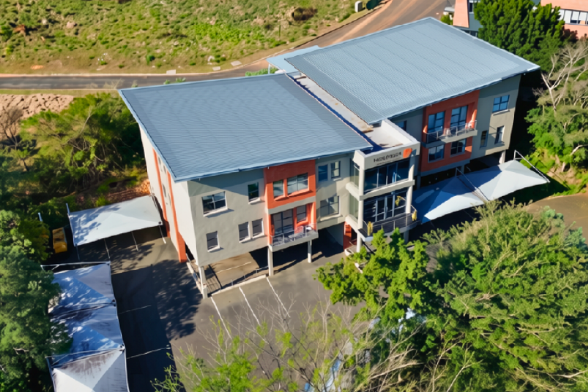 Aerial view of a multi-story commercial building featuring a metal roof, paved parking area with shade cloth carports, and surrounding landscaped grounds.