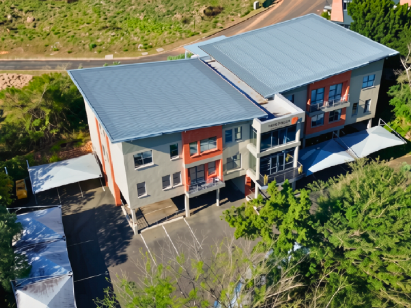 Aerial view of a multi-story commercial building featuring a metal roof, paved parking area with shade cloth carports, and surrounding landscaped grounds.
