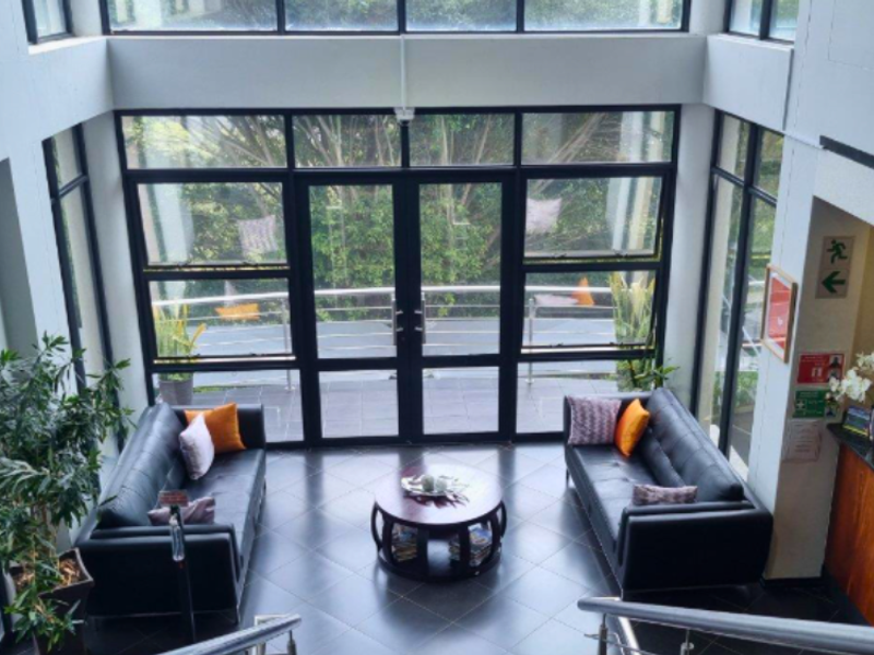 A bright, double-volume commercial entrance lobby featuring floor-to-ceiling glass windows, tiled flooring, and a professional reception area.