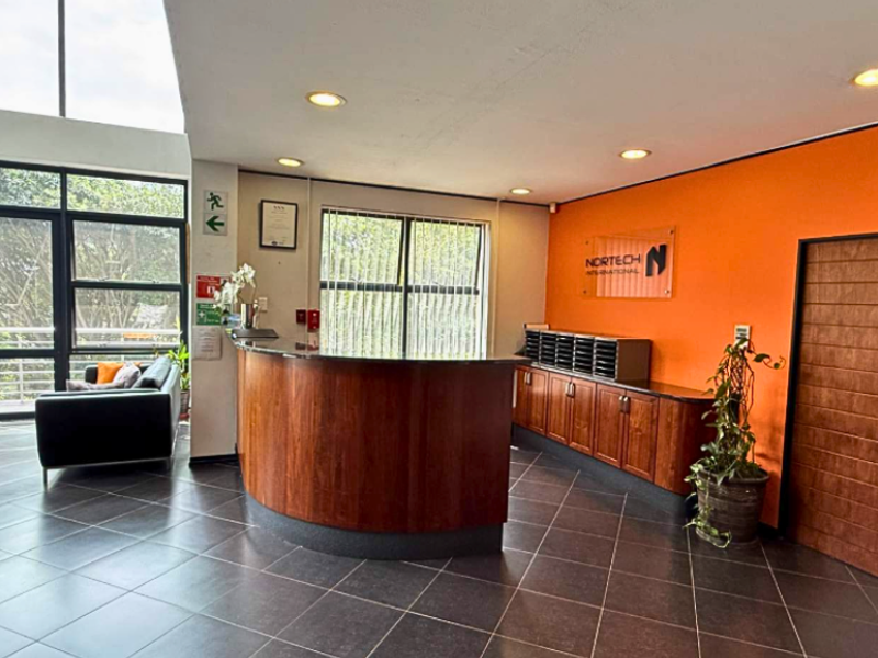 A bright commercial reception area featuring a curved wooden reception desk, built-in cabinetry, tiled flooring, and large windows providing natural light.