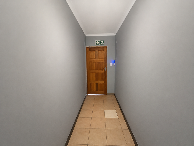 A clean, well-lit hallway featuring neutral grey walls, tiled flooring, and a solid wooden door with an emergency exit sign.
