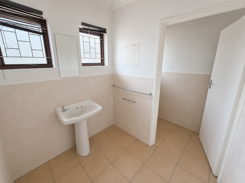 A clean, empty bathroom featuring a pedestal basin, tiled walls and flooring, and natural light from two windows with security bars.
