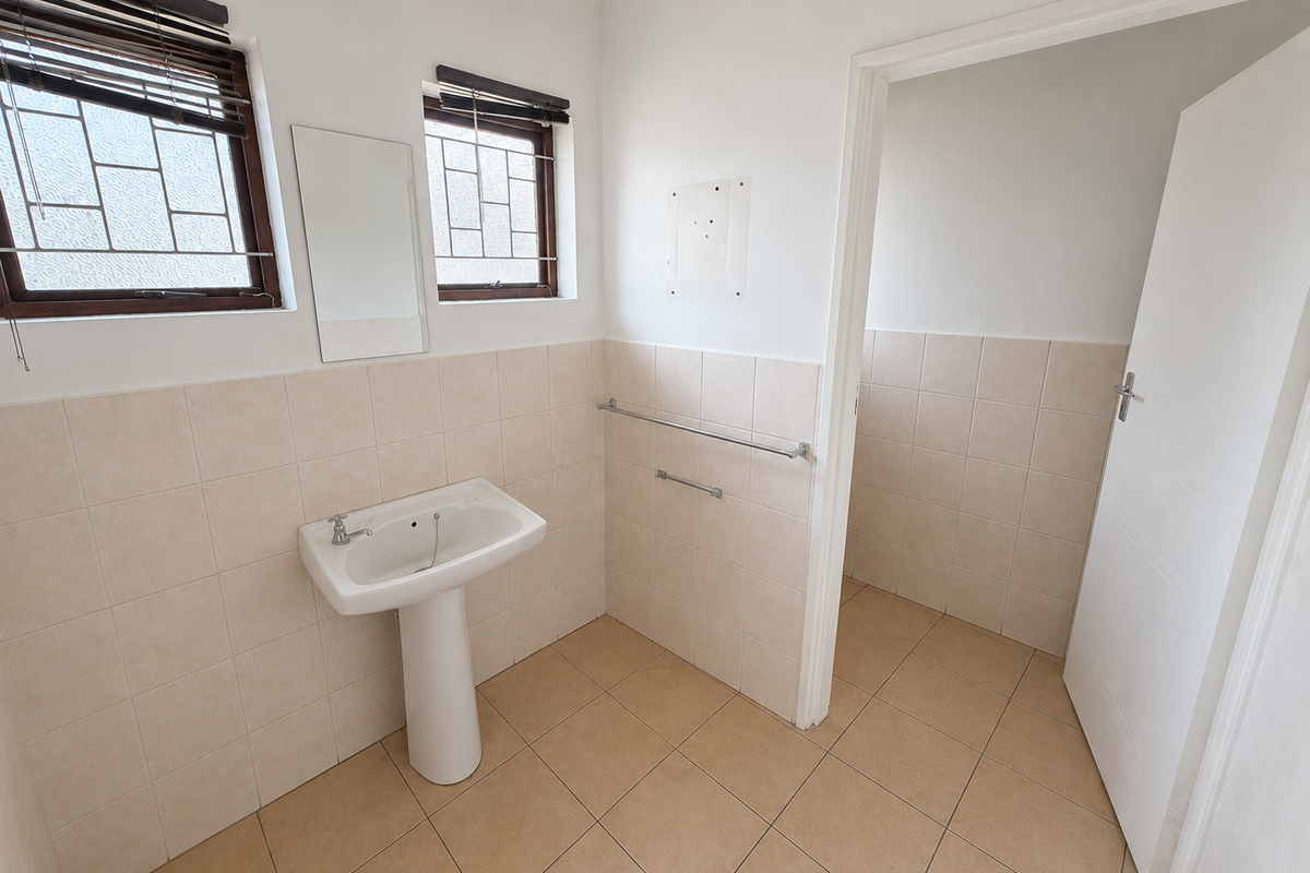 A clean, empty bathroom featuring a pedestal basin, tiled walls and flooring, and natural light from two windows with security bars.