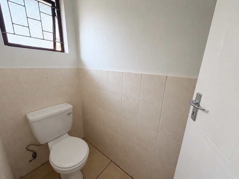 A compact, functional guest toilet featuring neutral ceramic wall and floor tiles, a standard white toilet suite, and a window with security bars.