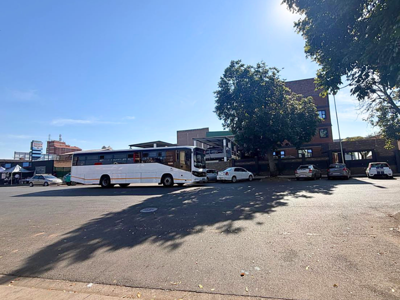 Street-level view of a commercial property featuring a multi-story brick building, paved parking area, and boundary wall.