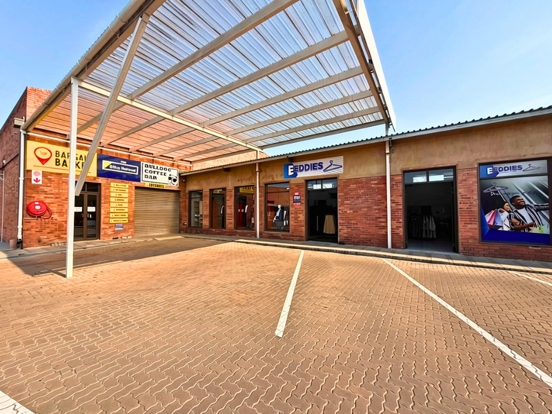 Commercial storefront complex featuring a paved parking area, covered walkway with a polycarbonate canopy, and multiple retail entrances with brick facade finishes.