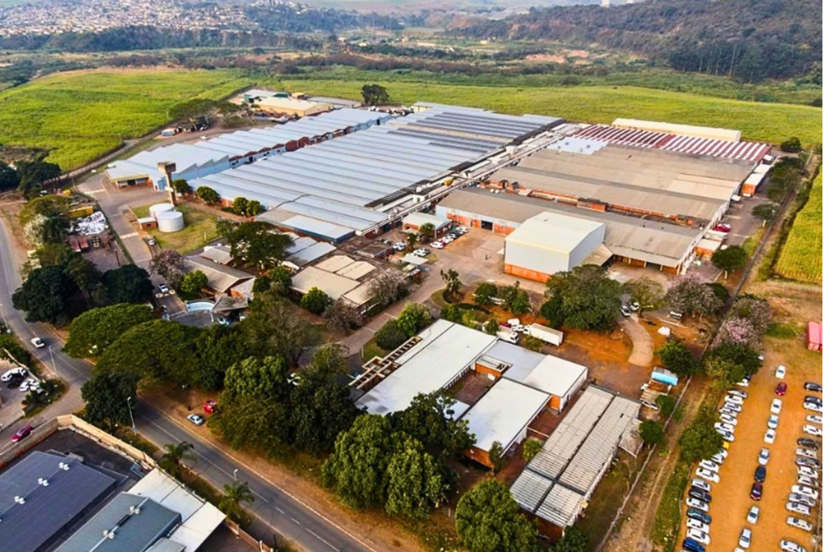 Aerial view of a large industrial facility featuring extensive warehouse structures, paved access roads, parking areas, and surrounding landscaped grounds.