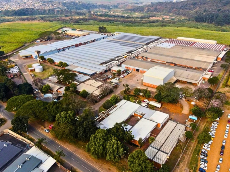 Aerial view of a large industrial facility featuring extensive warehouse structures, paved access roads, parking areas, and surrounding landscaped grounds.