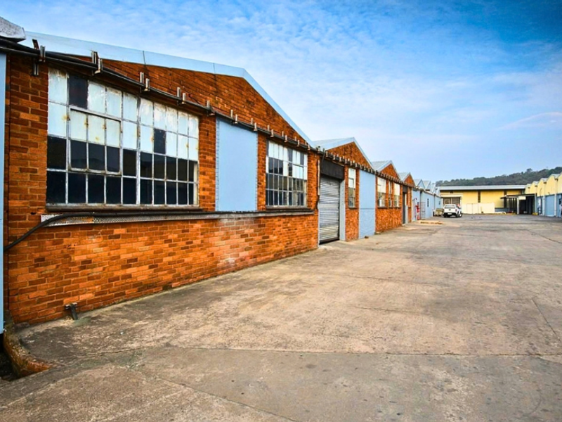 Exterior view of an industrial warehouse complex featuring brick facades, steel-framed windows, a roller shutter door, and a spacious concrete yard area.