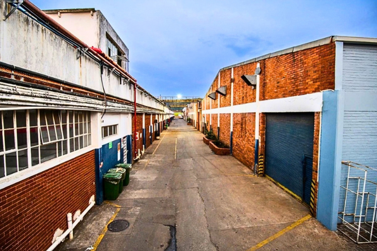 Industrial exterior view featuring a paved driveway between brick warehouse buildings with roller shutter access and steel-framed windows.