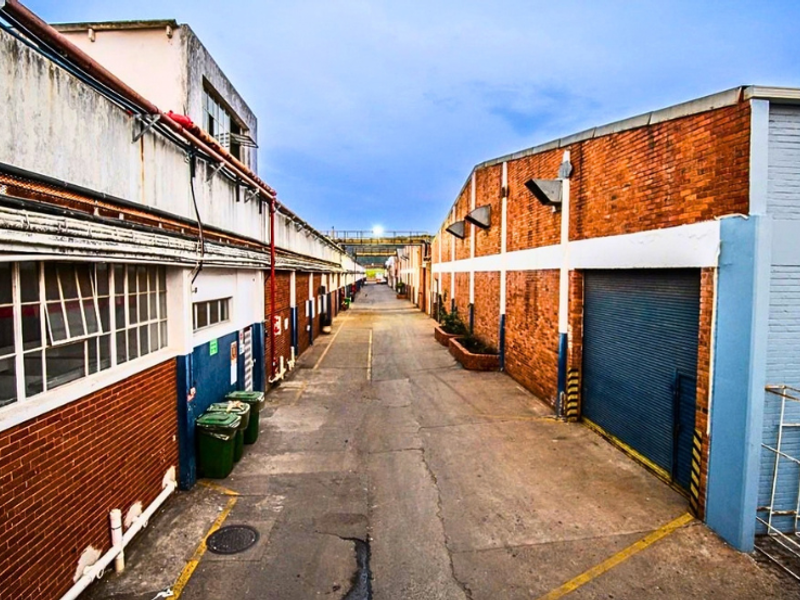 Industrial exterior view featuring a paved driveway between brick warehouse buildings with roller shutter access and steel-framed windows.