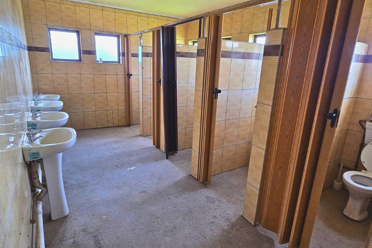 Industrial communal bathroom facility featuring multiple pedestal washbasins and private toilet cubicles with folding doors, finished with tiled walls and concrete flooring.