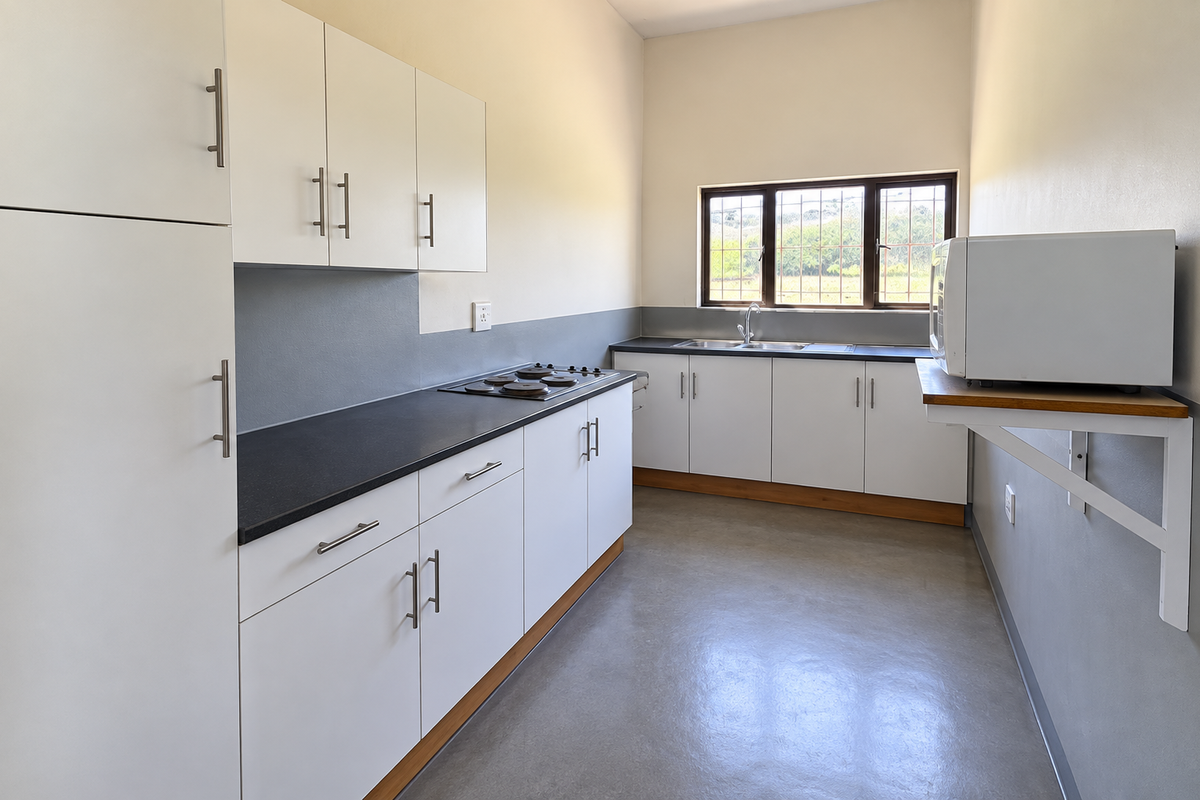 A clean, functional kitchen featuring white cabinetry, dark countertops, an integrated electric hob, stainless steel sink, and polished concrete flooring.
