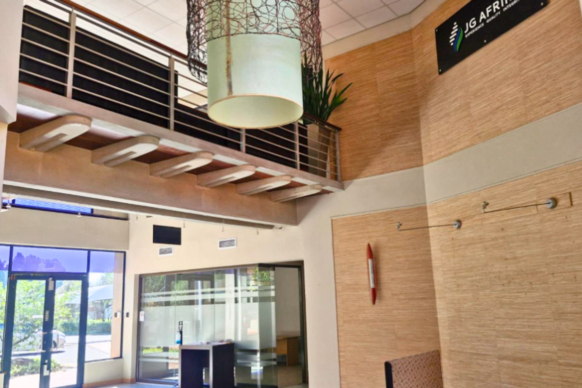 Spacious office reception area featuring a double-volume ceiling, mezzanine level with metal railing, glass partitions, and modern wood-look wall paneling.