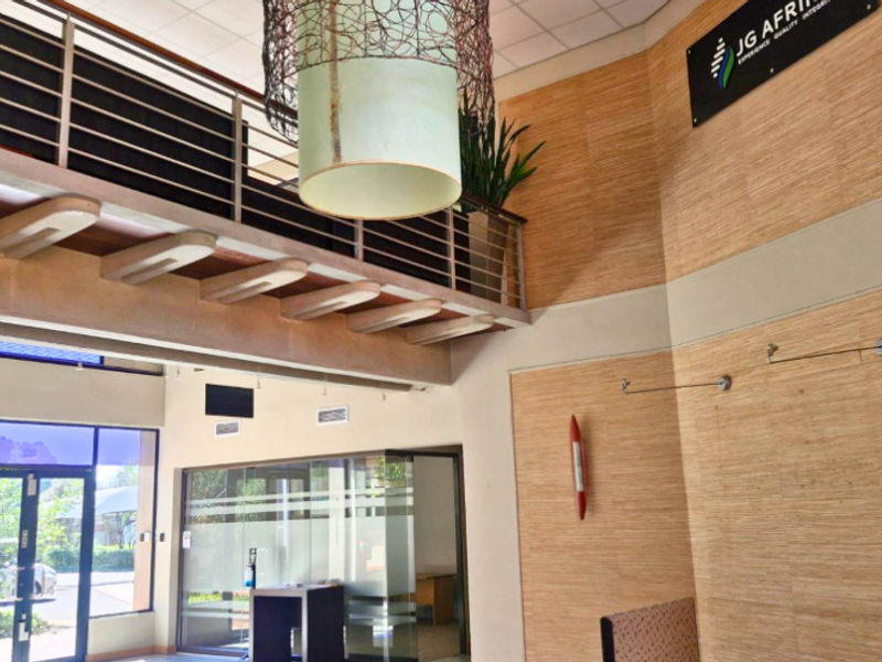 Spacious office reception area featuring a double-volume ceiling, mezzanine level with metal railing, glass partitions, and modern wood-look wall paneling.
