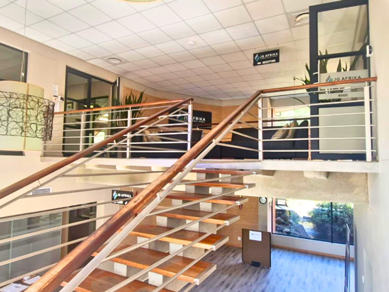 Modern office entrance featuring a striking open-tread wooden staircase with stainless steel balustrades, leading to an elevated mezzanine reception area.