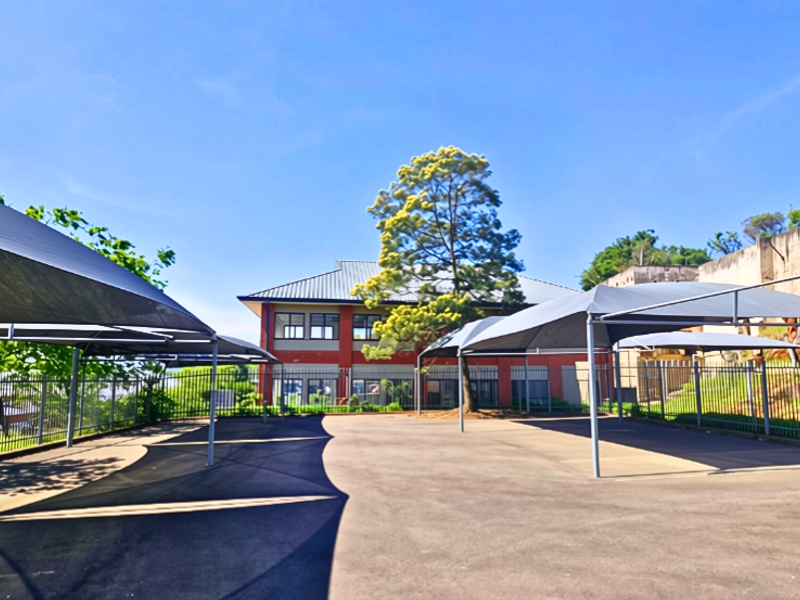 A paved office parking area featuring multiple shade cloth carports, enclosed by perimeter steel fencing with a brick office building in the background.