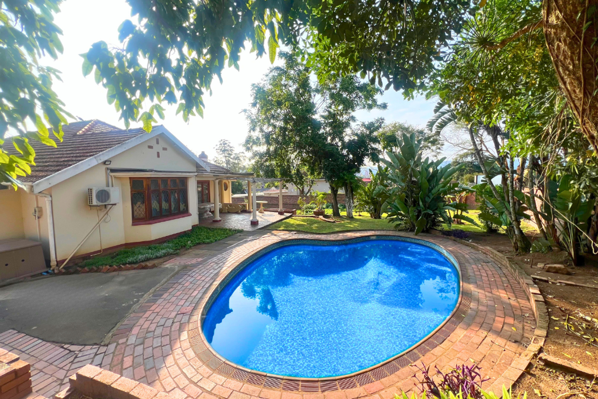 A rear exterior view featuring a kidney-shaped swimming pool with brick surround, a covered patio area, and established garden landscaping.