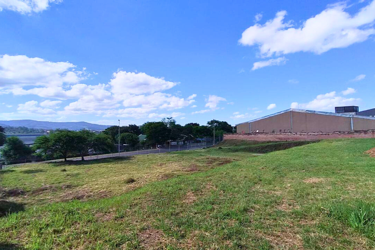 An expansive, undeveloped plot of land featuring open grass areas, perimeter fencing, and views of the surrounding industrial landscape and distant hills.