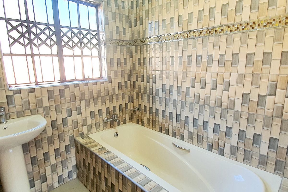 A functional bathroom featuring a pedestal basin, a built-in bathtub with tiled surround, and a window with security bars.