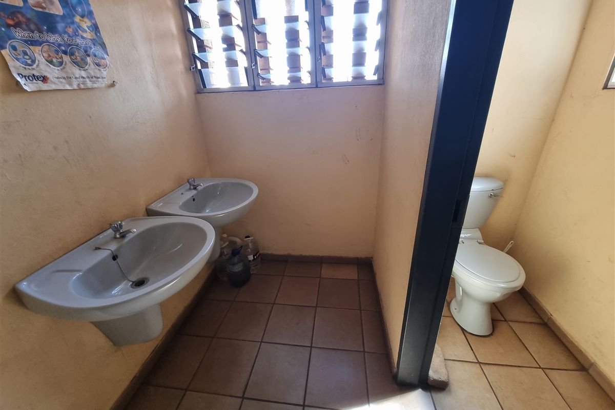 A functional office bathroom featuring two pedestal wash basins, tiled flooring, and a separate toilet cubicle with louvered windows for ventilation.