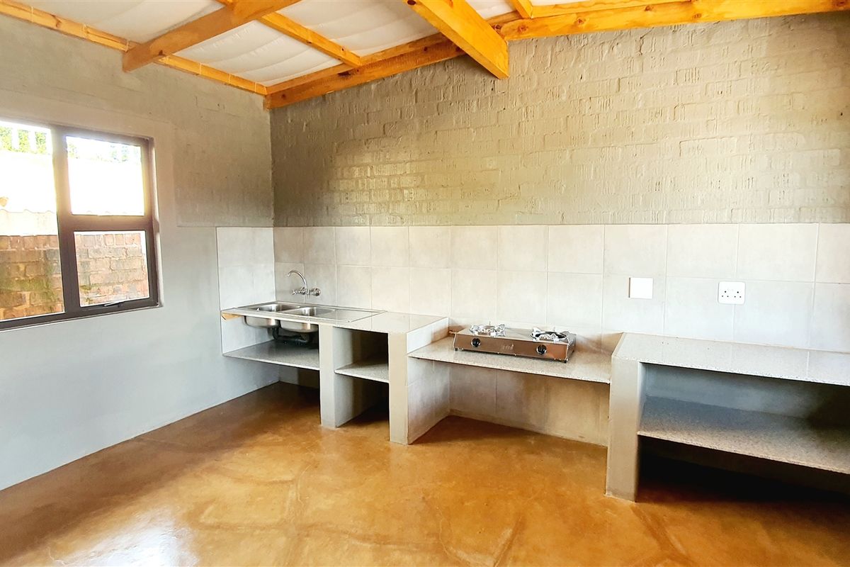 A rustic, unfinished kitchen area featuring concrete countertops, a double basin sink, exposed brick walls, and timber ceiling beams.