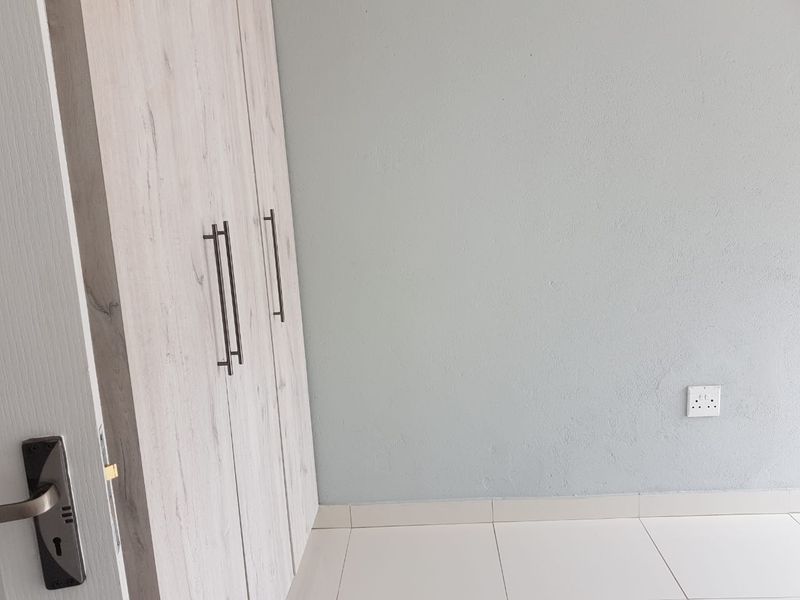 A clean, empty bedroom featuring light wood-finish built-in cupboards, neutral painted walls, and modern white floor tiles.