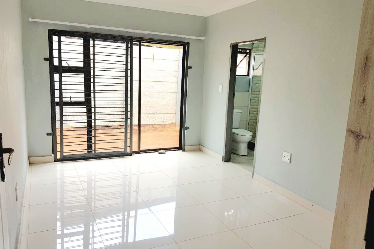 A bright, empty bedroom featuring polished tile flooring, a sliding glass door with security gate, and direct access to an en-suite bathroom.