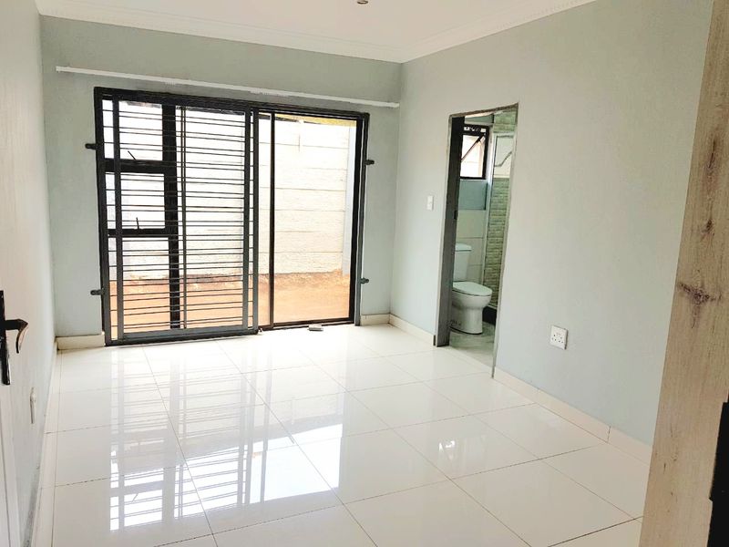 A bright, empty bedroom featuring polished tile flooring, a sliding glass door with security gate, and direct access to an en-suite bathroom.