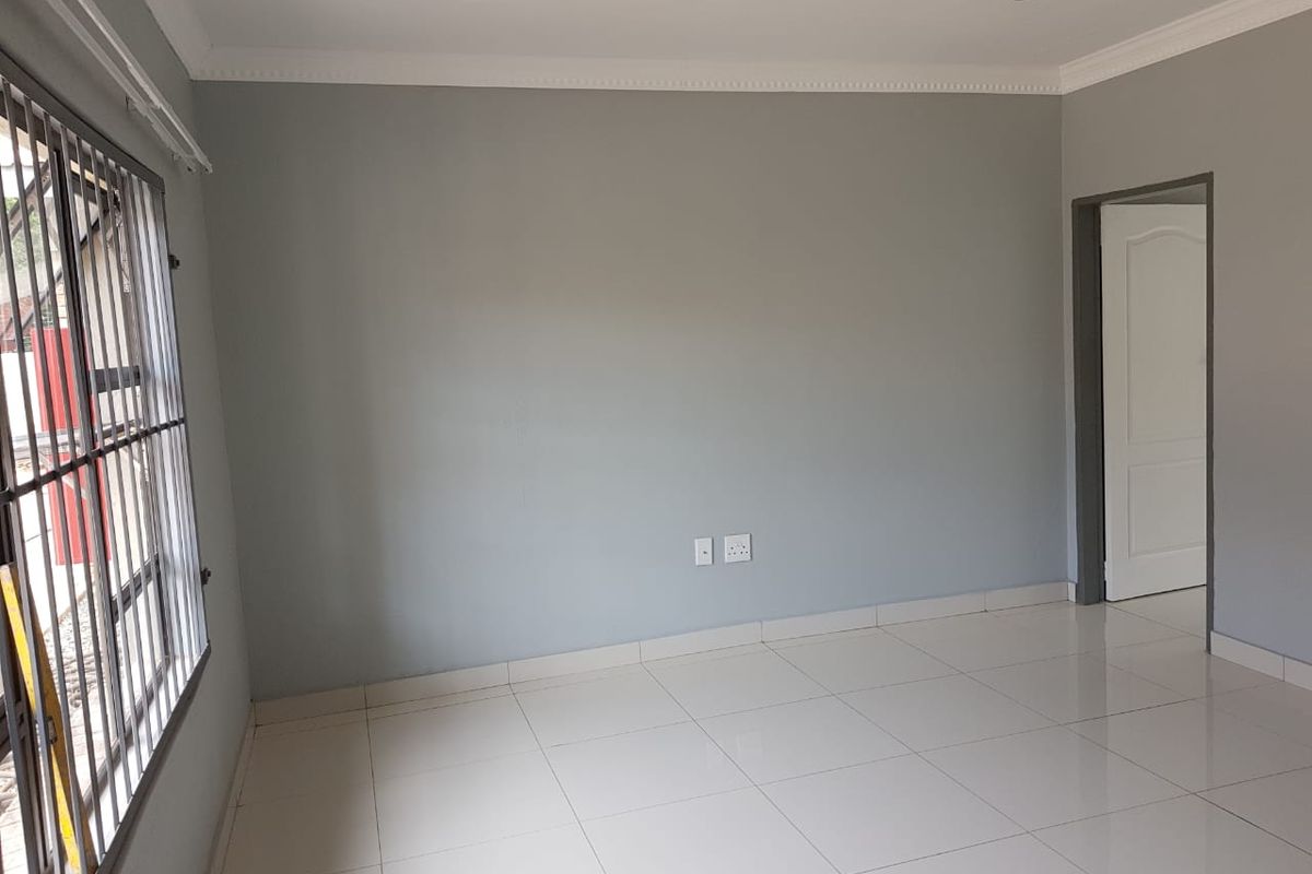 A bright, empty living room featuring neutral grey walls, white tiled flooring, recessed downlighting, and security bars on the window.