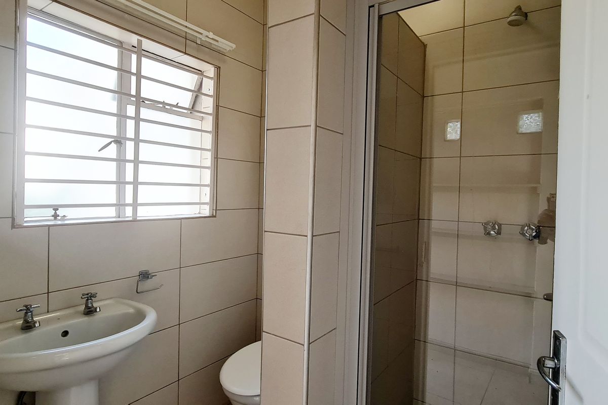 A functional bathroom featuring a pedestal washbasin, toilet, and a separate shower enclosure with tiled walls and a window with security bars.