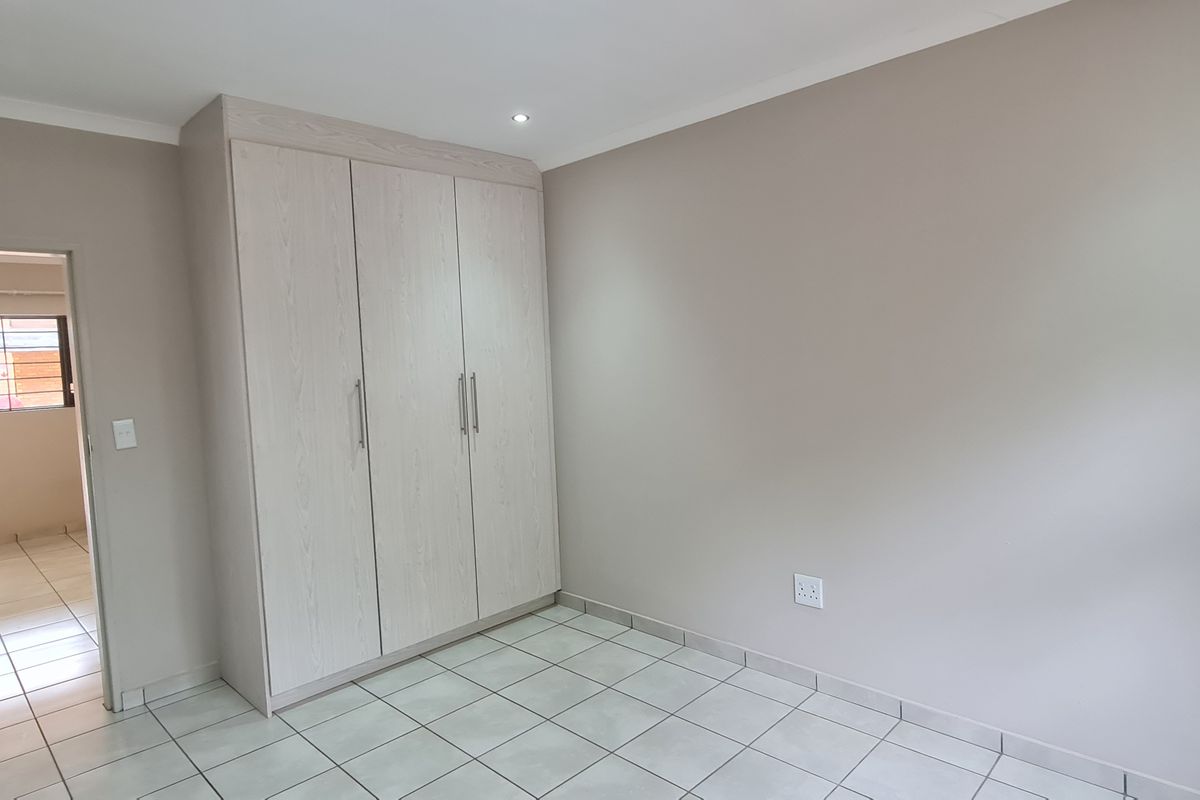 A clean, neutral bedroom featuring built-in cupboards, tiled flooring, and recessed ceiling lighting, with a view into an adjacent tiled room.