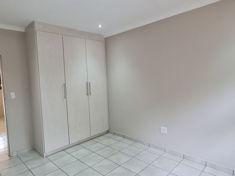 A clean, neutral bedroom featuring built-in cupboards, tiled flooring, and recessed ceiling lighting, with a view into an adjacent tiled room.