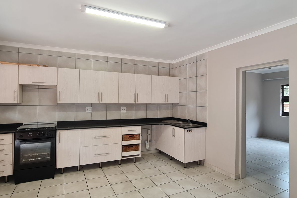 A functional kitchen featuring light wood-grain cabinetry, black countertops, tiled splashbacks, and a freestanding oven, with an adjacent room visible through the doorway.