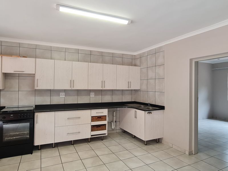 A functional kitchen featuring light wood-grain cabinetry, black countertops, tiled splashbacks, and a freestanding oven, with an adjacent room visible through the doorway.