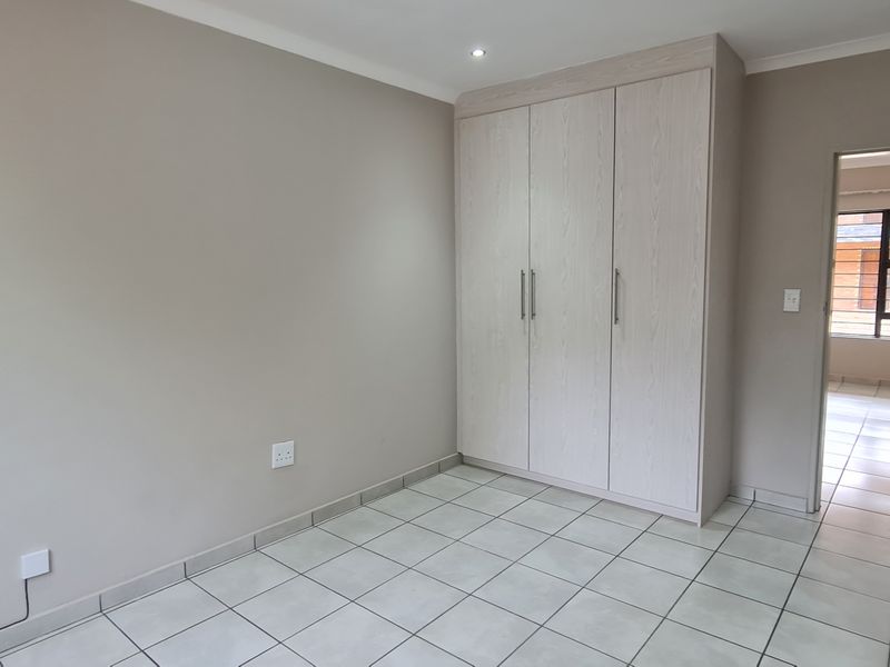 A bright, empty bedroom featuring neutral wall tones, tiled flooring, and built-in light wood-grain cabinetry, with a view into the adjacent hallway.