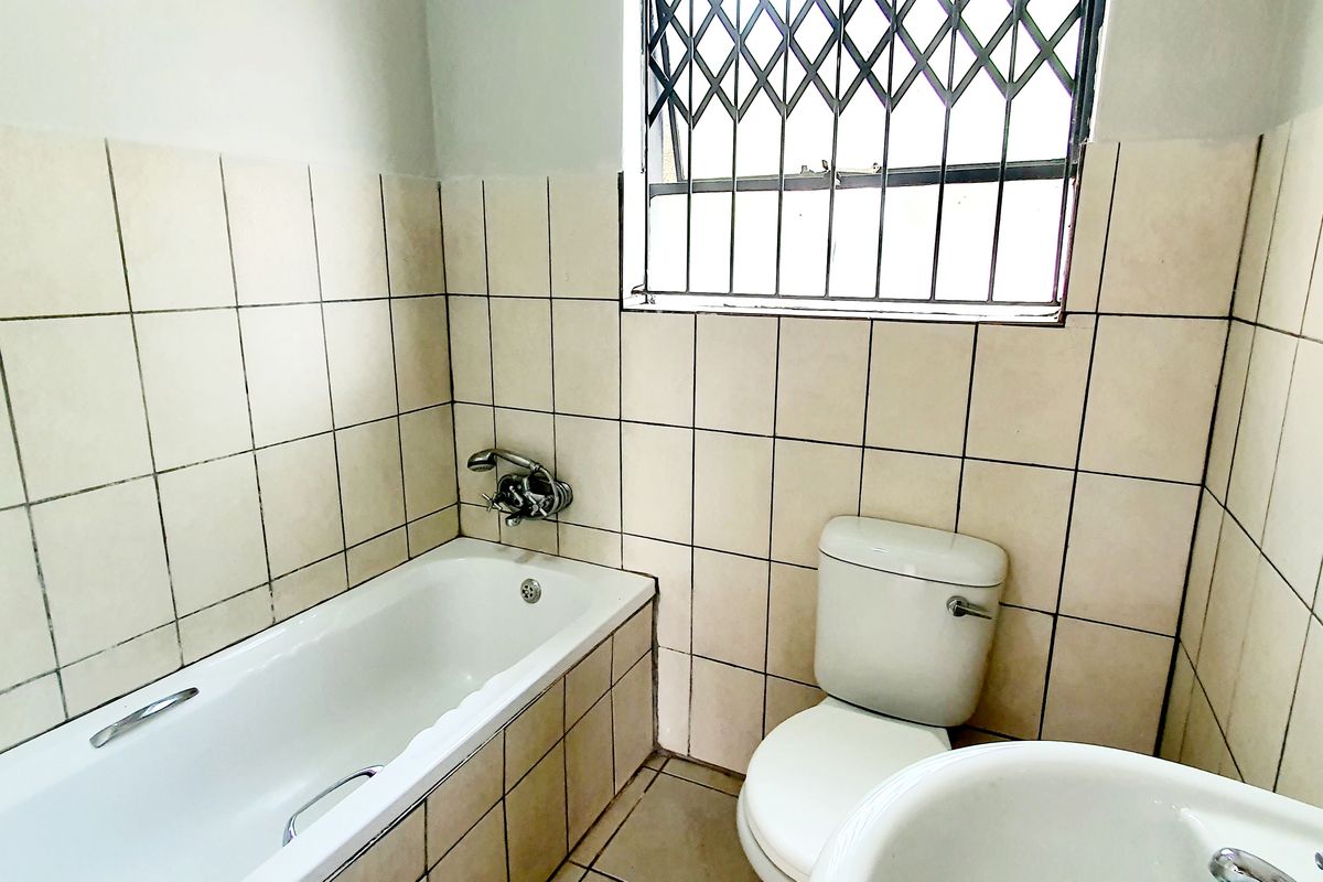 A functional bathroom featuring a standard bathtub, toilet, and pedestal basin with tiled walls and a security-fitted window.