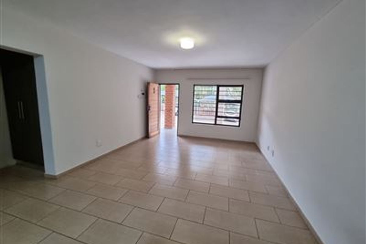 A spacious, empty living area featuring neutral tiled flooring, clean white walls, and natural light through an aluminium-framed window.