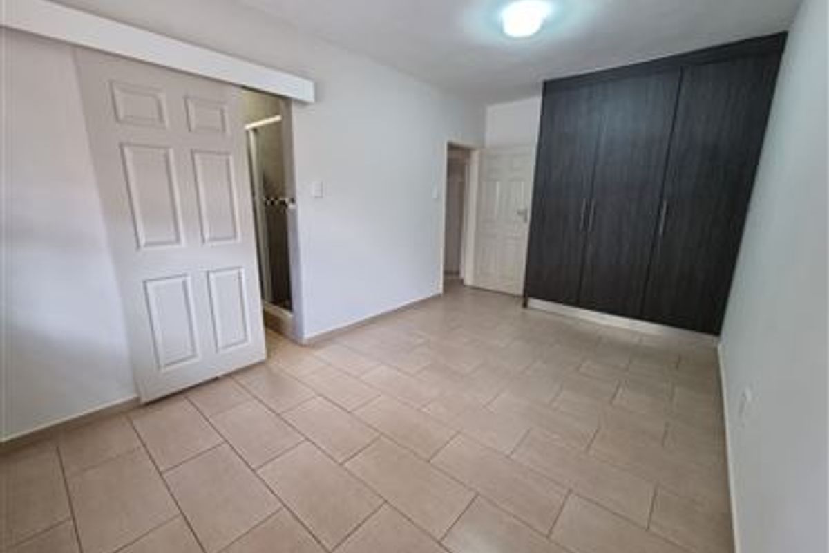 A clean, empty bedroom featuring neutral tiled flooring, dark wood-finish built-in cupboards, and a sliding door leading to an en-suite bathroom.