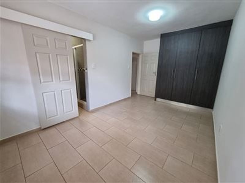 A clean, empty bedroom featuring neutral tiled flooring, dark wood-finish built-in cupboards, and a sliding door leading to an en-suite bathroom.