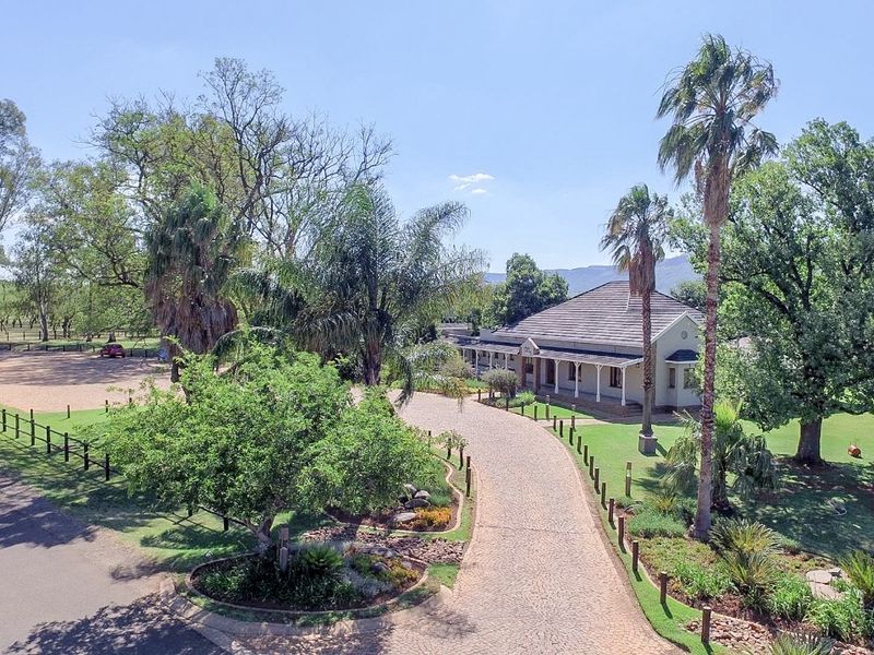 Aerial view of a residential property featuring a long cobblestone driveway, mature landscaped gardens, and a well-maintained dwelling with a covered veranda.