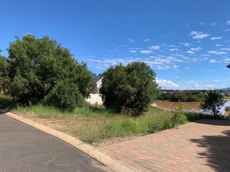 A vacant residential plot featuring a paved driveway, asphalt road access, and established trees with a view of the nearby water body.