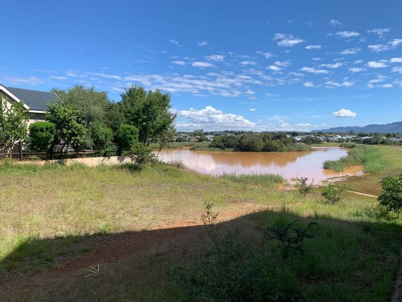 A scenic view of a natural lake surrounded by lush greenery and distant residential landscape under a clear blue sky.