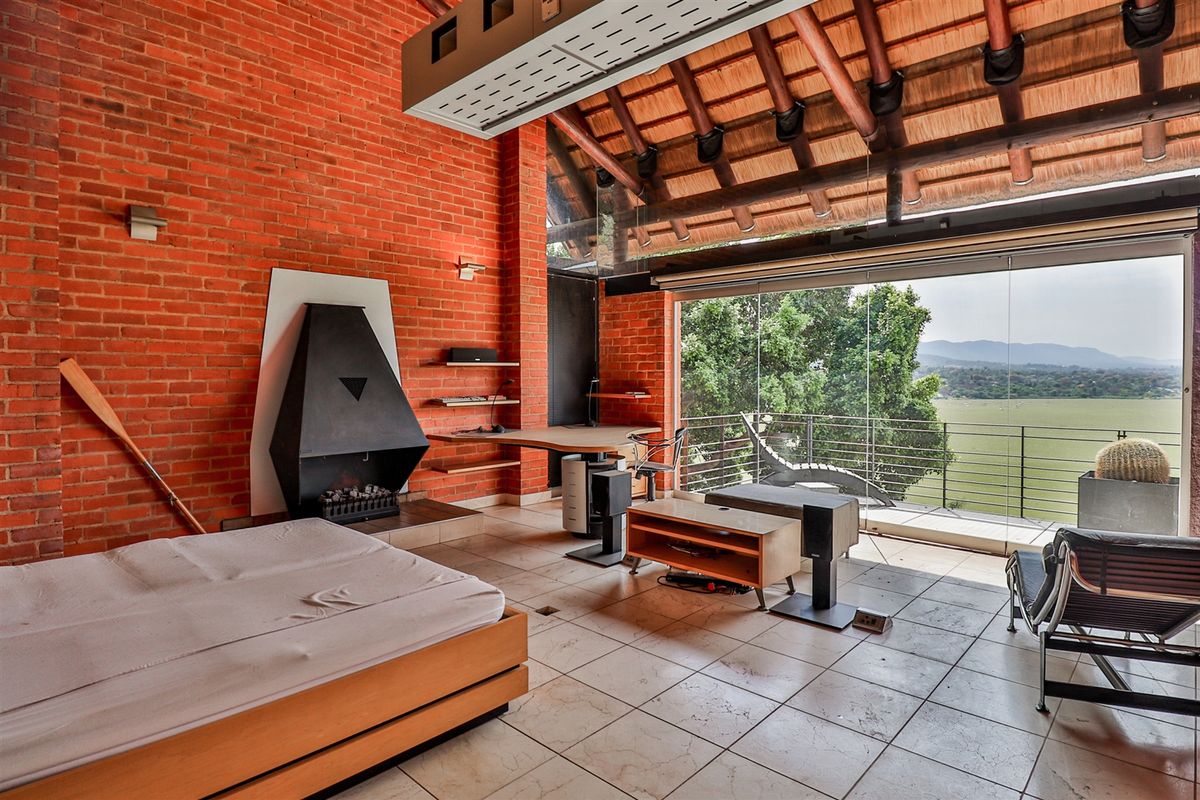 Spacious bedroom featuring exposed brick walls, a high thatched ceiling with timber beams, a built-in fireplace, and expansive glass doors opening to a scenic view.
