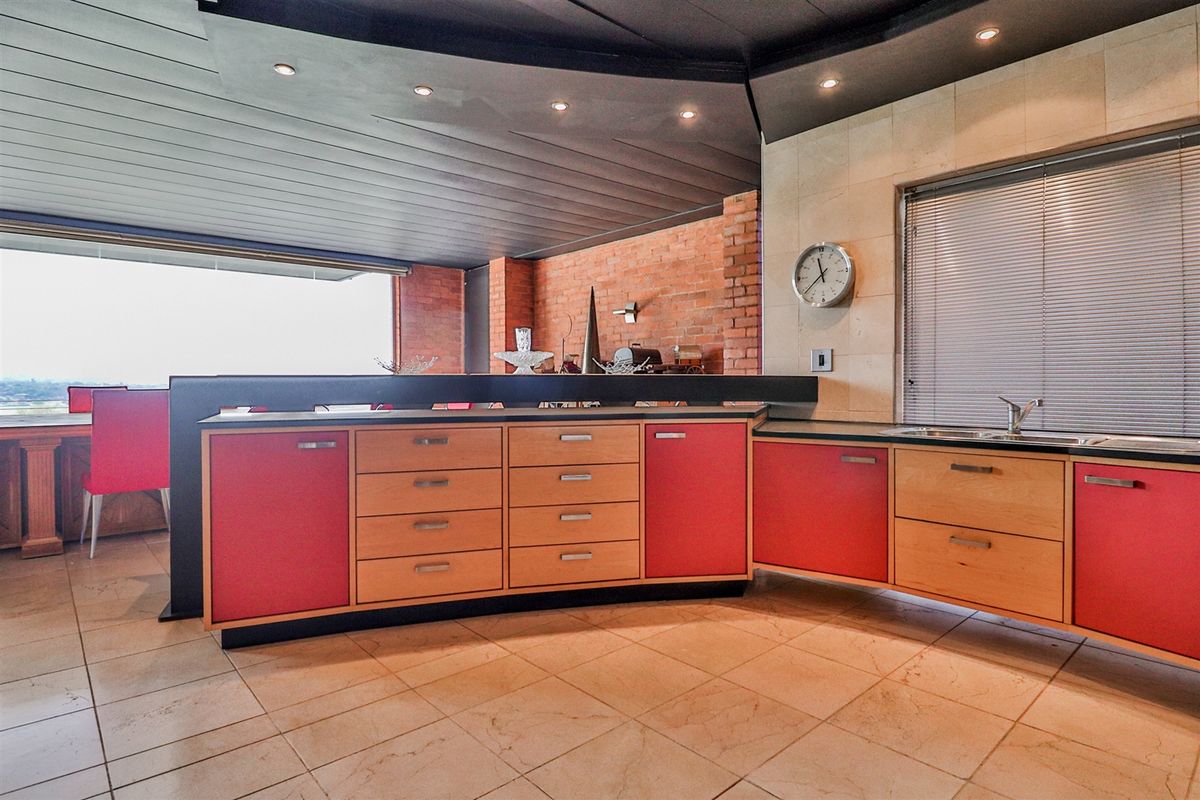 A contemporary kitchen featuring red and light wood cabinetry, stone tile flooring, an exposed brick feature wall, and a breakfast bar overlooking the living area.